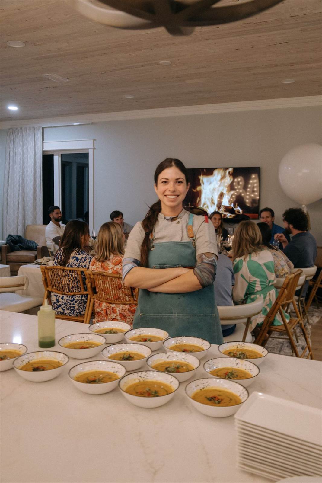 Chef in an apron standing behind rows of soup bowls with a dinner party in the background