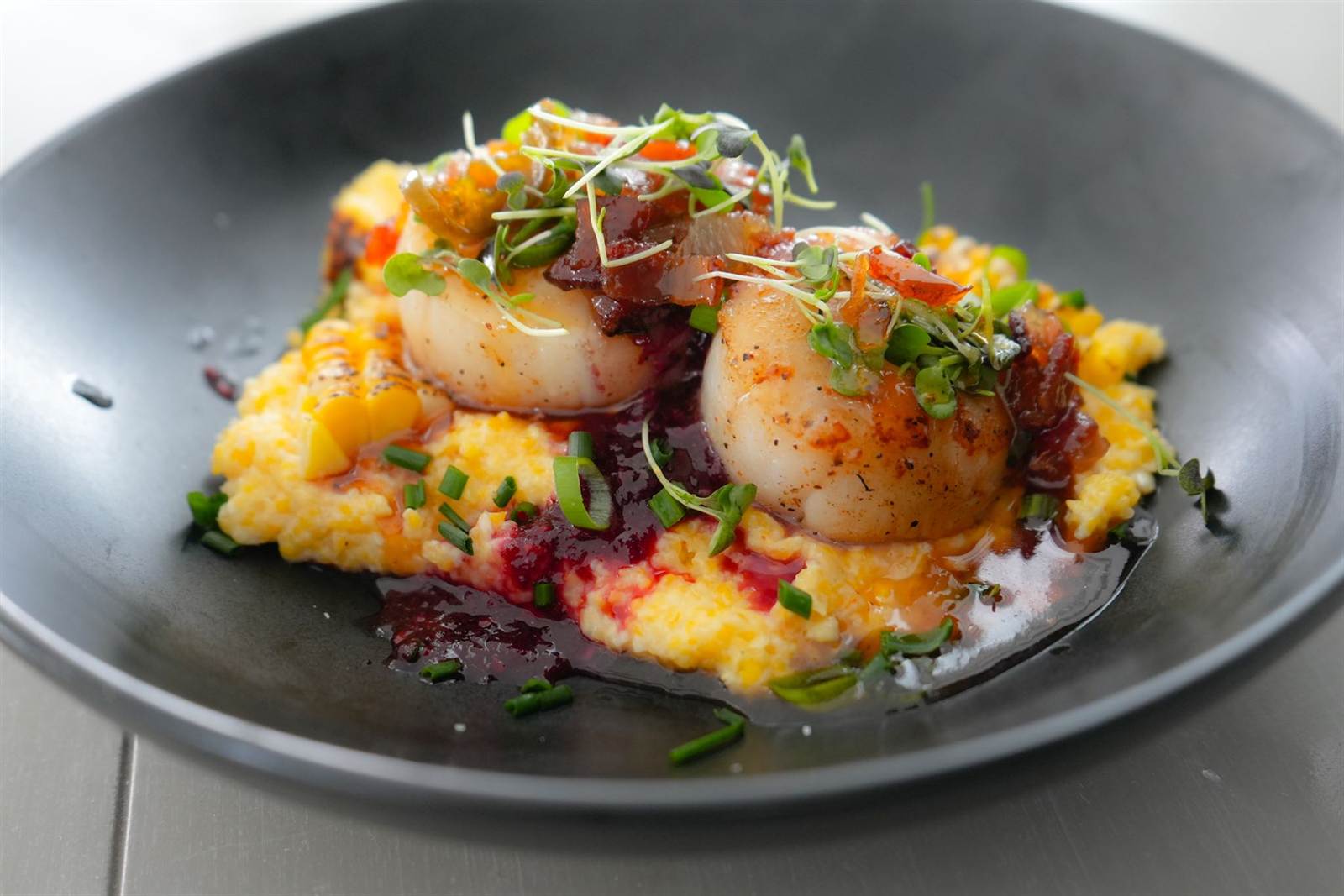Seared scallops over gouda grits with micro greens in a dark bowl